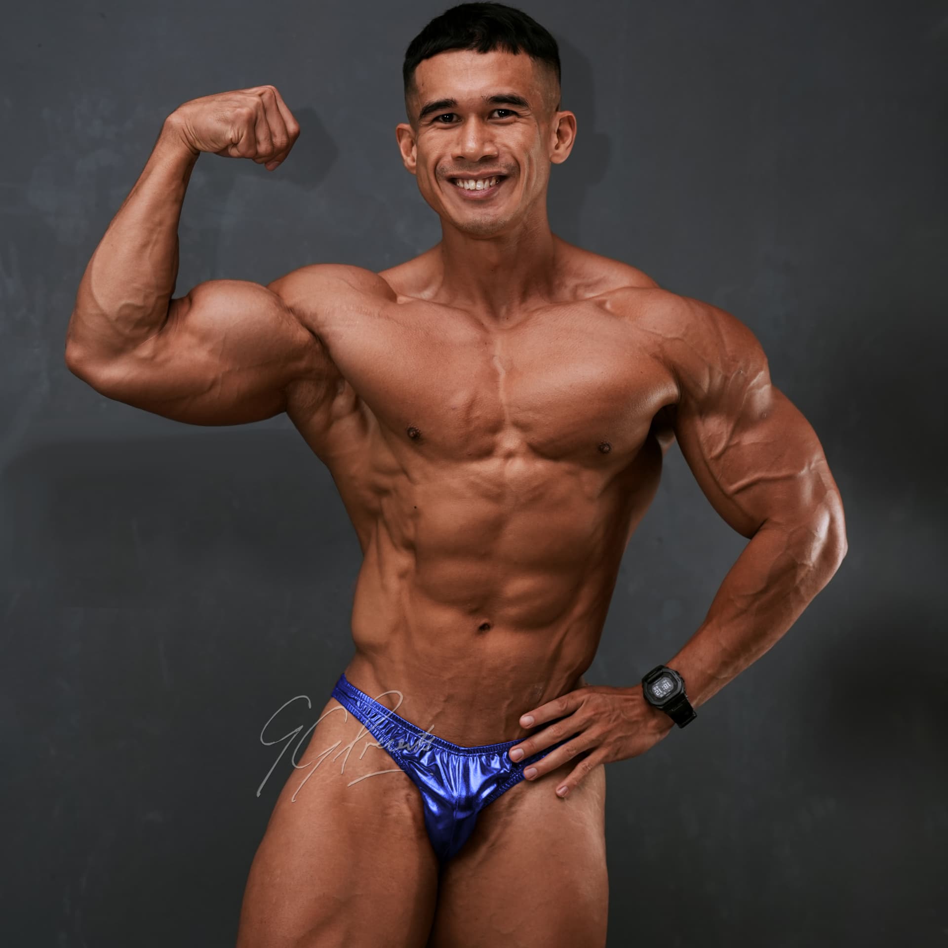 Athlete flexing biceps in blue posing trunks against a grey studio backdrop