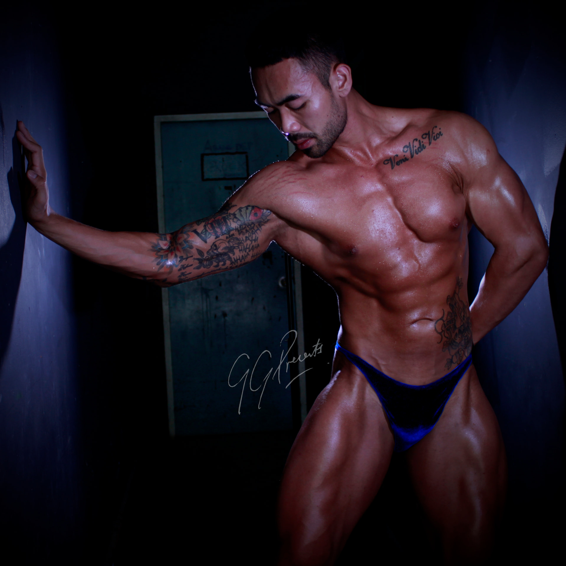 Tattooed athlete in blue posing trunks leaning against a dark alley wall