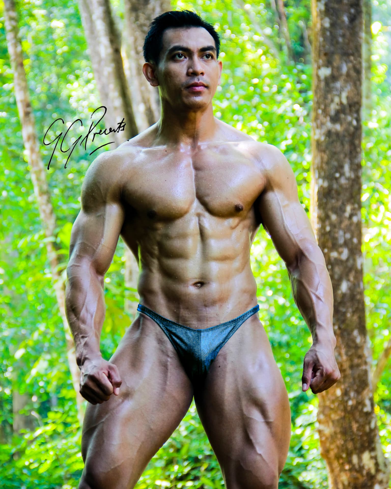 Muscular athlete standing in a lush green jungle wearing blue posing trunks